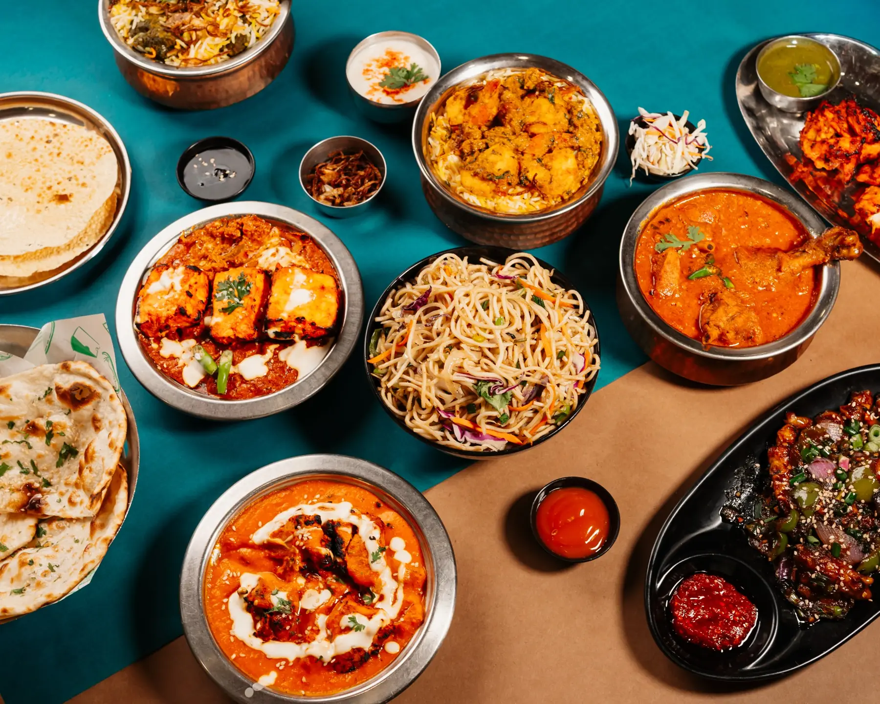 A spread of Park View dishes on teal and kraft paper: biryani, paneer tikka masala, noodles, chilli chicken, curries and breads